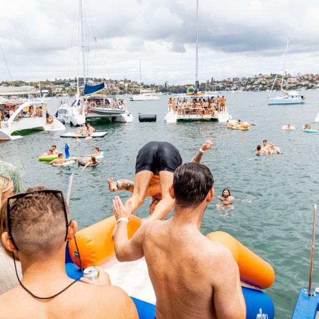 People gather on boats and in the water at a lake or marina for a social event; one person dives into the water from an inflatable platform, while others watch and swim. Consider Sydney boat party hire for your next gathering to enhance the experience with added fun and excitement.