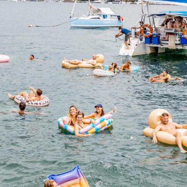 People are floating on inflatable rafts and swimming in a marina, with boats and a cityscape in the background. In the distance, a catamaran party Sydney adds to the lively atmosphere.