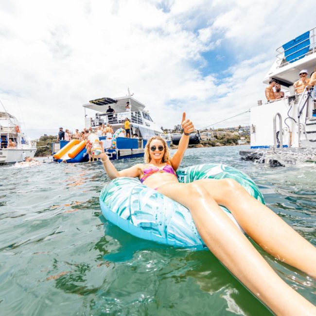 A person lounges on an inflatable raft in the water, giving a thumbs-up, with several boats and other people in the background enjoying a sunny day. It's the perfect scene for a Sydney boat party hire.