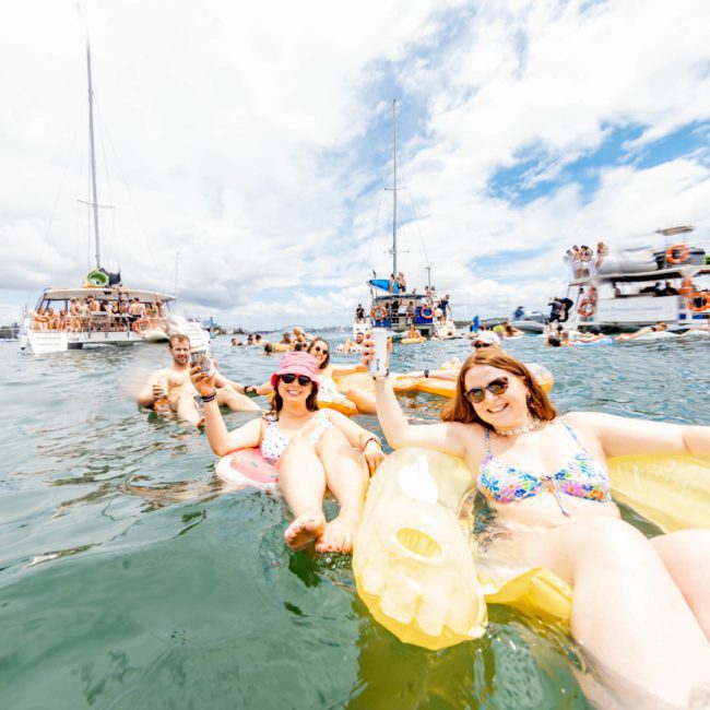 A lively scene with people on inflatables in the water, smiling and holding drinks, with boats and more people in the background on a sunny day. It's a perfect moment for a Sydney boat party hire or even a private yacht charter on Sydney Harbour.