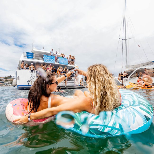 Two people in inflatable pool tubes relax in the water, raising drinks towards each other, with several boats and many people in the background; it's a perfect day for a Sydney boat party hire.