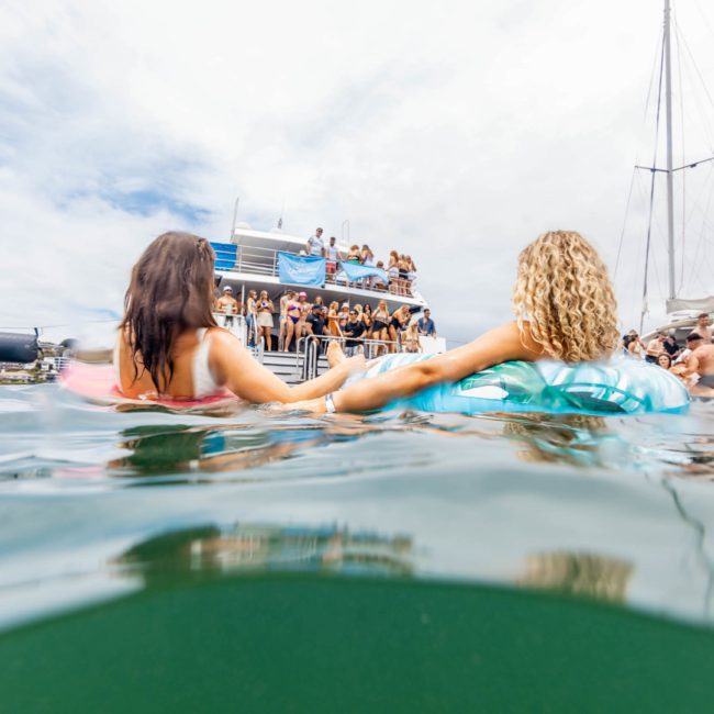 Two people on inflatable tubes hold hands while floating in water near a private yacht charter on Sydney Harbour under a partly cloudy sky.