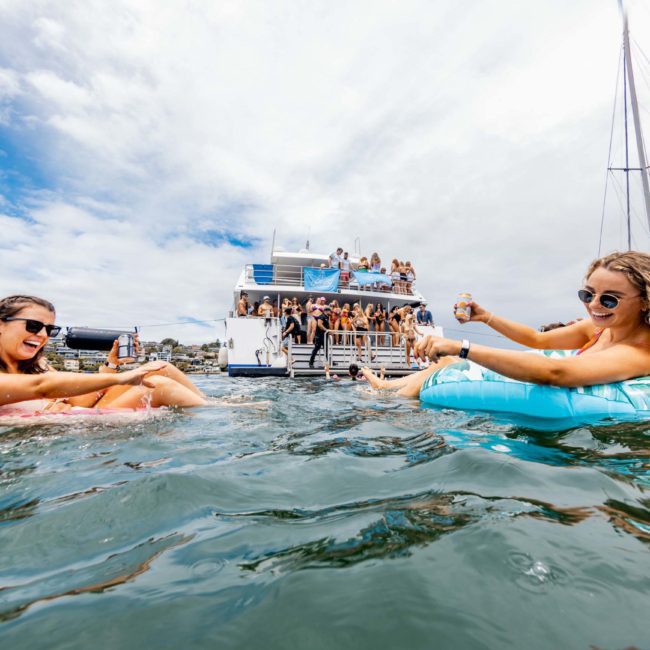 Two women float leisurely in the water while toasting with drinks, enjoying a luxury yacht hire Sydney, with people on a nearby boat and a sailboat in the background.