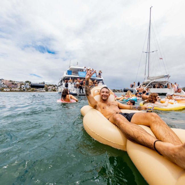 A man relaxes on an inflatable float in the water with boats and other people in the background on a cloudy day, possibly enjoying a luxury yacht hire Sydney experience.