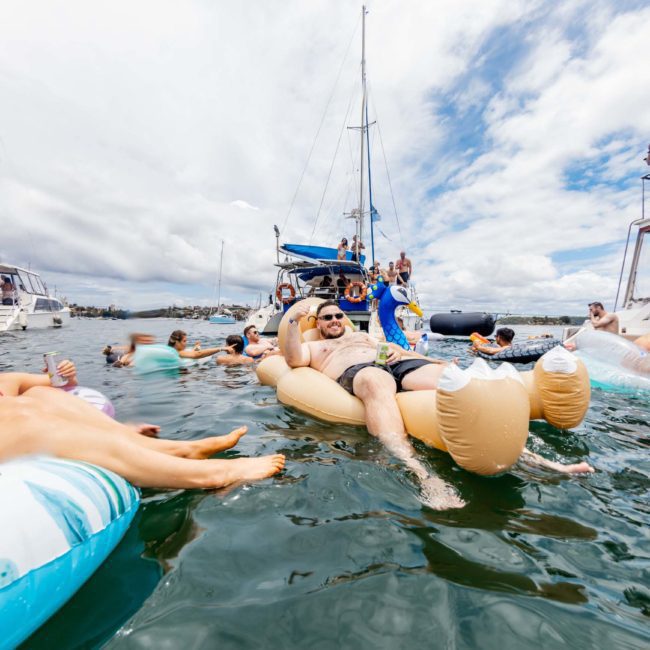 People on inflatable floats and boats enjoy a sunny day on the water, with some lounging and others engaging in activities. Various boats, including luxury yacht hires from Sydney, are anchored nearby, creating a festive atmosphere.