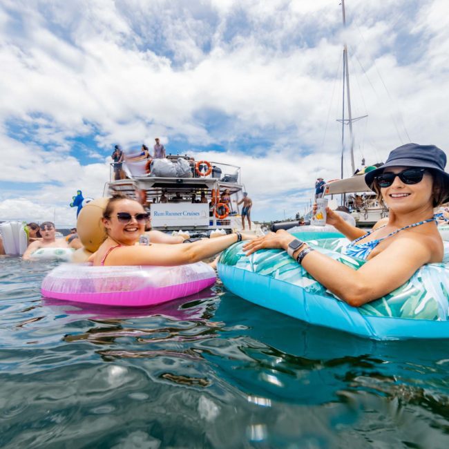 Two people float in inflatable tubes in the water, smiling at the camera. Several other people and boats are in the background under a partly cloudy sky, highlighting the fun of a Catamaran party Sydney event.