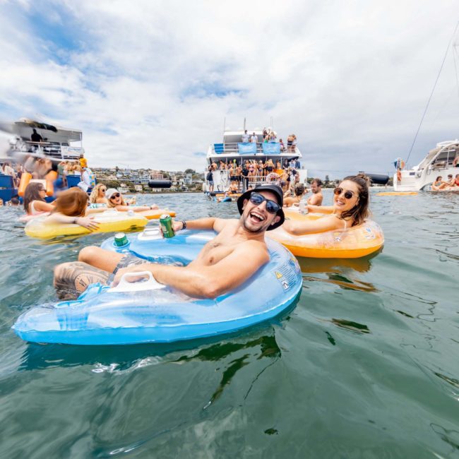 People enjoy a sunny day on inflatable floats and boats in the water. A man in sunglasses smiles and holds a drink, while others relax around him. Boats and more people are visible in the background, showcasing the delights of luxury yacht hire Sydney Harbour.