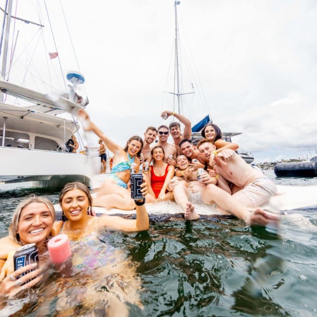 A group of people enjoying themselves on a floating platform in the water, with several boats anchored in the background, likely part of a private yacht charter Sydney Harbour.