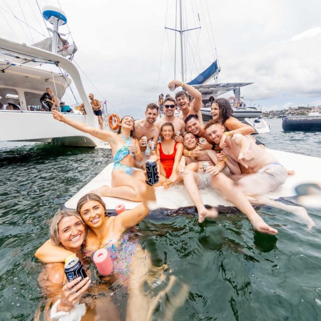 A group of people in swimsuits enjoy drinks and pose for a photo on a small floating mat in the water, with boats visible in the background, perfect for capturing moments at corporate boat events Sydney.