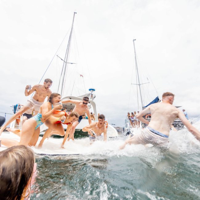 A group of people in swimwear are having fun on a boat, with one person jumping into the water while others look on. Several boats are anchored in the background, showcasing the excitement of a Sydney boat party hire.