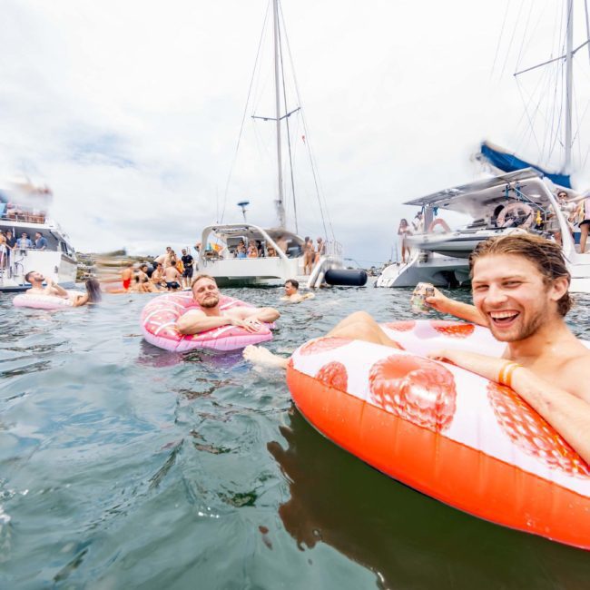 A group is enjoying a day on the water with inflatable floats, surrounded by several boats. The sky is cloudy, and some people are swimming while others relax on the floats. It's a lively scene, resembling a delightful Sydney boat party hire or even a Catamaran party in Sydney.
