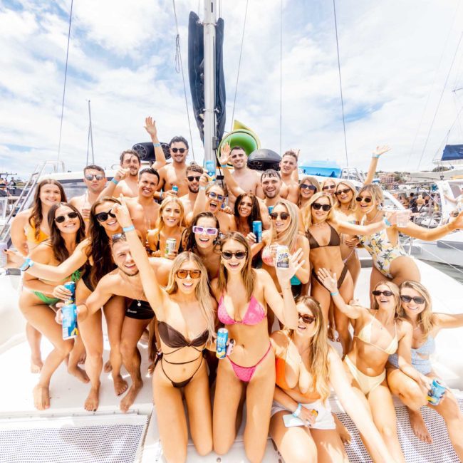 A large group of people in swimsuits and sunglasses are gathered on the deck of a luxury yacht hire in Sydney, smiling and holding drinks. The sky is clear with a few clouds in the background.