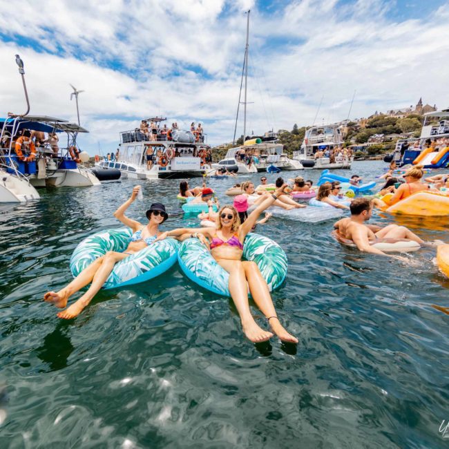 People are floating on inflatable pool floats in the water near several anchored boats, creating a lively scene under the blue sky. Many are enjoying a Catamaran party Sydney, making the most of this vibrant day by the water.