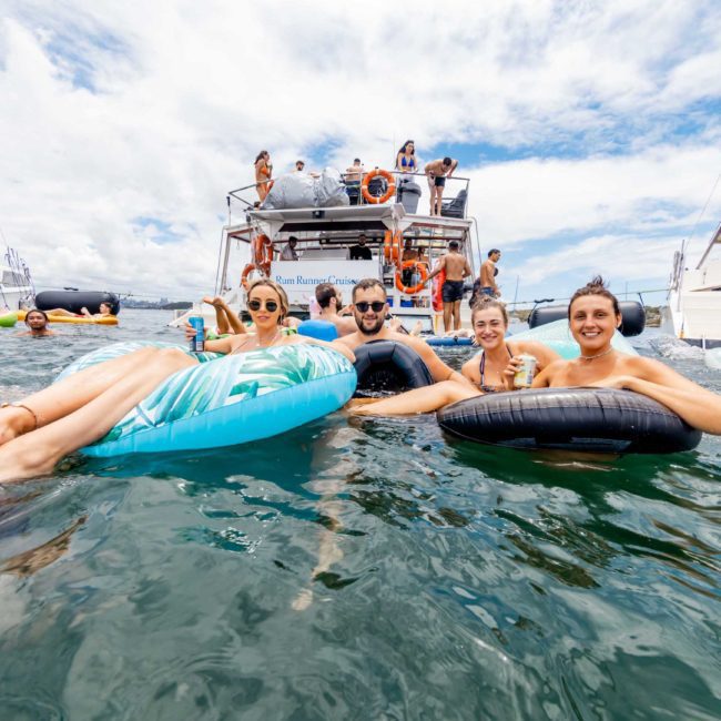 A group of people relax on inflatable pool floats in the water near a boat on a sunny day, enjoying what seems to be a corporate boat event in Sydney. Additional boats and individuals are visible in the background.