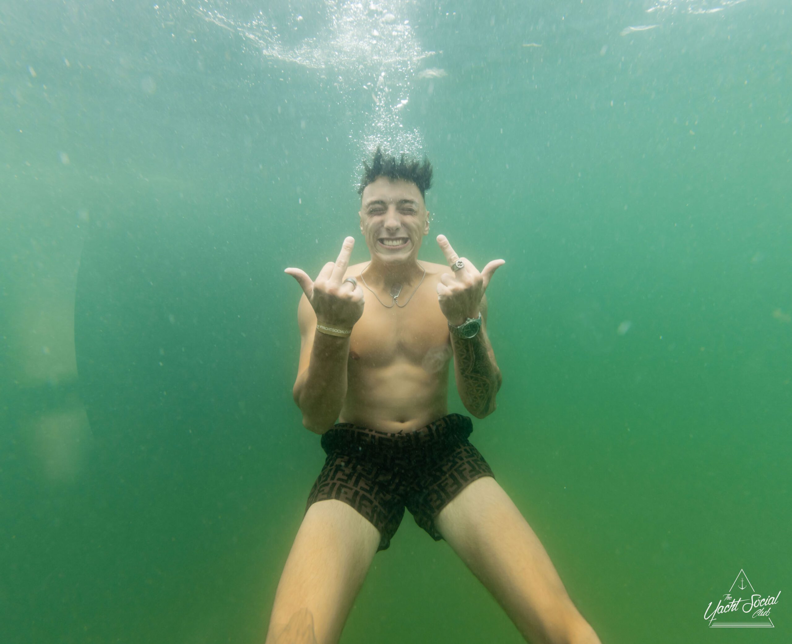 A person underwater wearing black shorts, smiling, and holding up two middle fingers while celebrating a Sydney boat party hire.