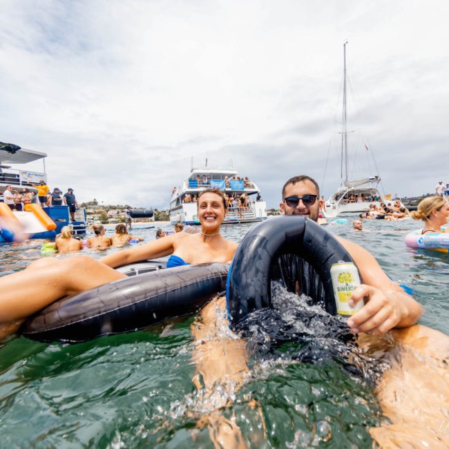 Two people relax on pool floats with drinks in hand, surrounded by others on various inflatables and boats, including a DJ boat hire Sydney, in the background on a partly cloudy day.