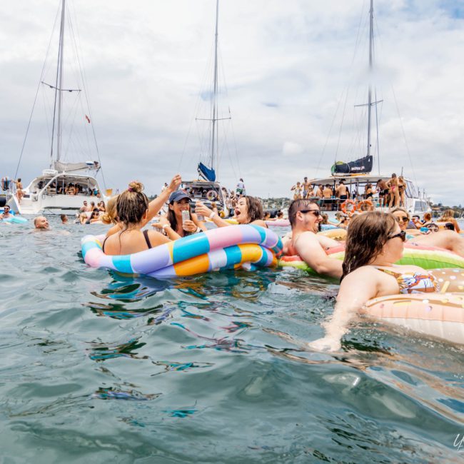 People are floating on inflatable rafts and swim rings in the water near boats on a cloudy day, enjoying the serene atmosphere of a luxury yacht hire Sydney.