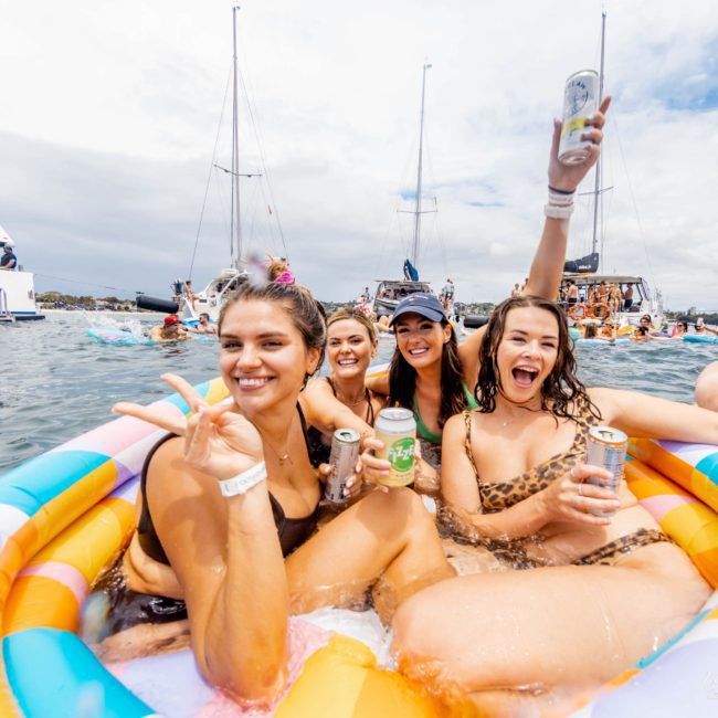 A group of four women in swimsuits enjoying drinks while sitting in an inflatable pool on the water, with boats and other partygoers in the background. It's a vibe reminiscent of a catamaran party Sydney style.