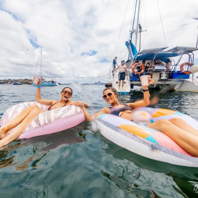 Two people in swimsuits float on inflatable rings holding drinks near a private yacht charter Sydney Harbour, with several others on board, in a scenic, sunny water setting.