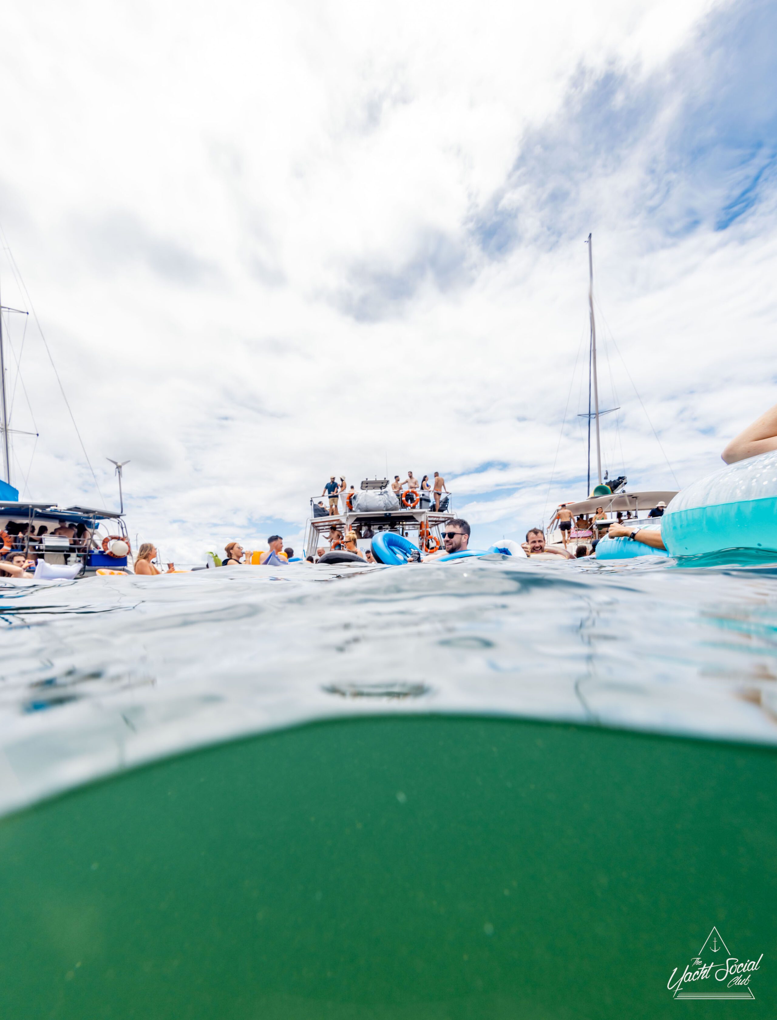 A group of people enjoy recreational activities on and around boats in a body of water under a partly cloudy sky, taking advantage of private yacht charter options in Sydney Harbour.