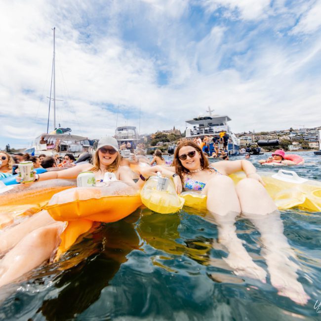 A group of people floating on inflatables in the water, holding drinks and smiling. Several boats, including a luxury yacht hire Sydney, are anchored in the background under a partly cloudy sky.