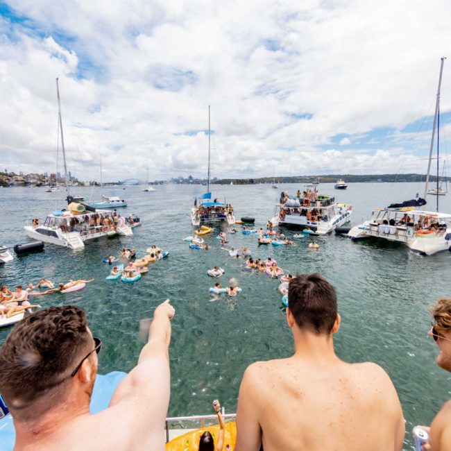 A group of people on a boat point towards others swimming and lounging on floaties near other boats in a sunny, crowded bay scene. Ideal for luxury yacht hire Sydney, this vibrant setting promises a memorable day on the water.
