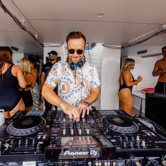A DJ in a patterned shirt mixes music on a Pioneer DJ console at an event aboard a catamaran. People in swimsuits are visible in the background, setting the perfect mood for this lively catamaran party in Sydney.