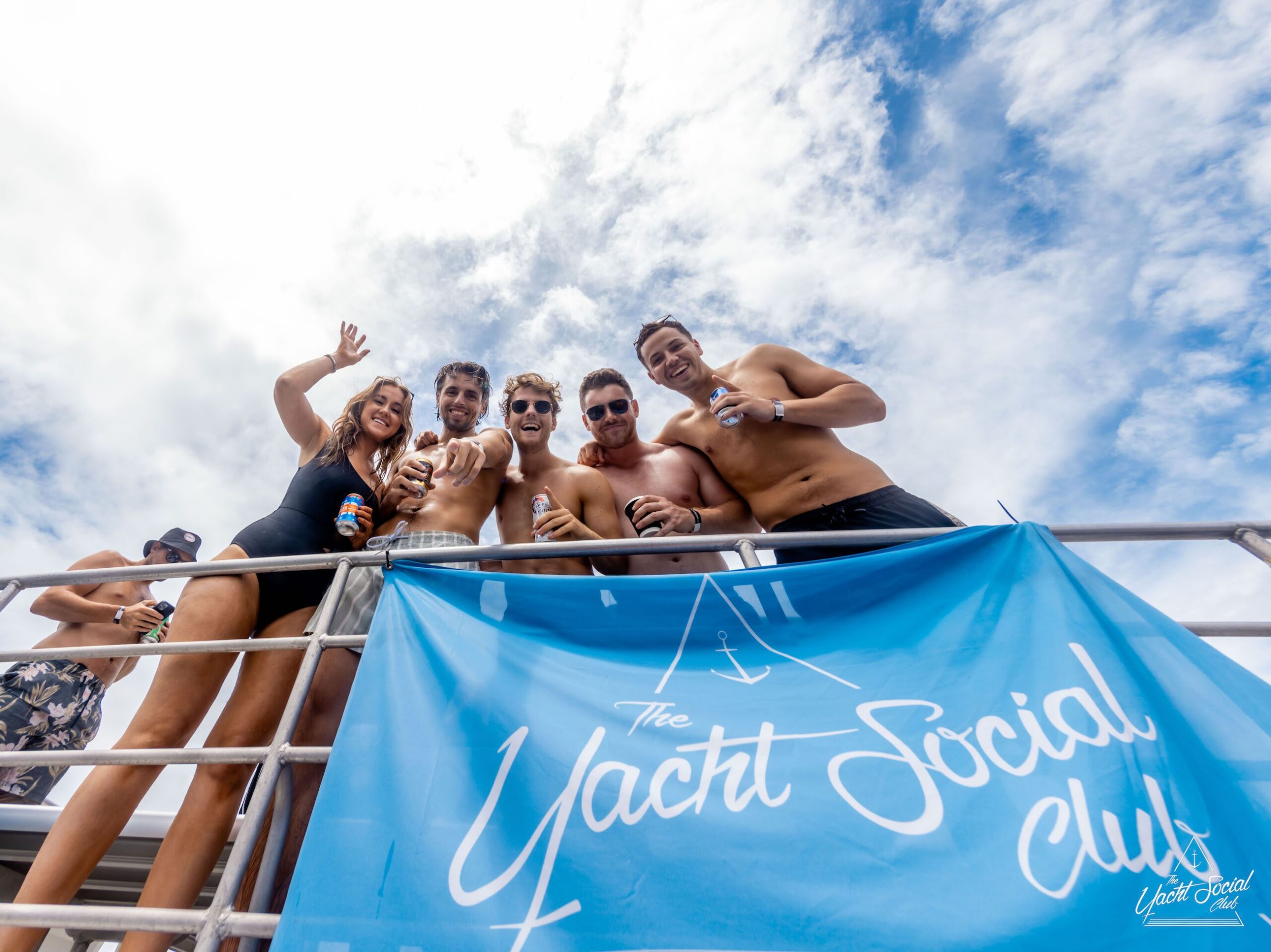 A group of people in swimwear stands on a yacht holding drinks, with a "Yacht Social Club" banner displayed below them against a partly cloudy sky backdrop, enjoying the ultimate Sydney boat party hire.