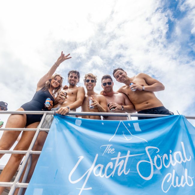 A group of five people stands on a boat, smiling and holding drinks. A blue banner reading "The Yacht Social Club" hangs below them against a partly cloudy sky. Enjoy the ultimate Sydney boat party hire for a memorable day on the water.