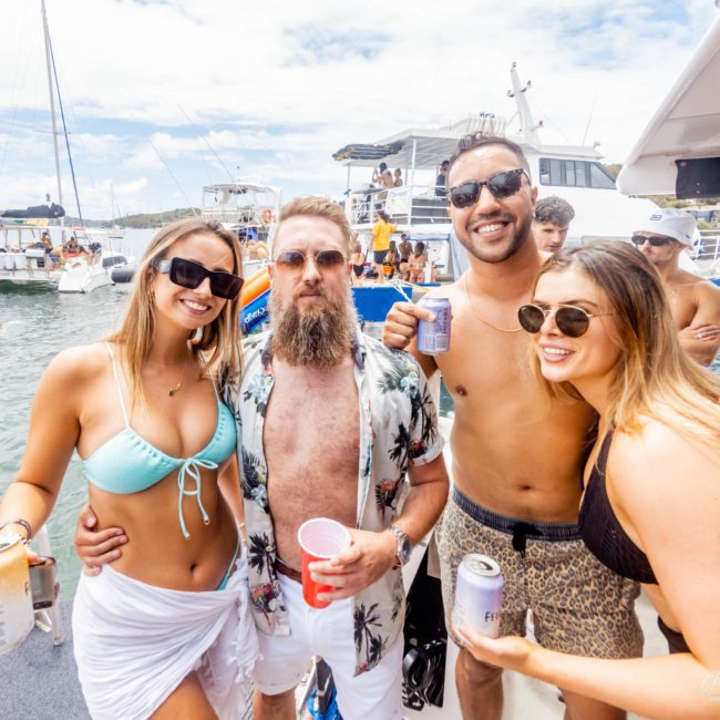 Four people in swimsuits, two men and two women, pose together on a boat with drinks in their hands at a lively Sydney boat party hire. Other boats and people are visible in the background on the water, creating a festive atmosphere.