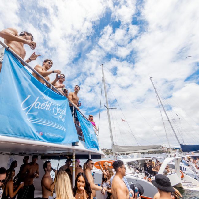 A group of people stand and socialize on a docked yacht at a Private yacht charter Sydney Harbour boat party on a sunny day. Other boats and people are in the background, enjoying the vibrant atmosphere.