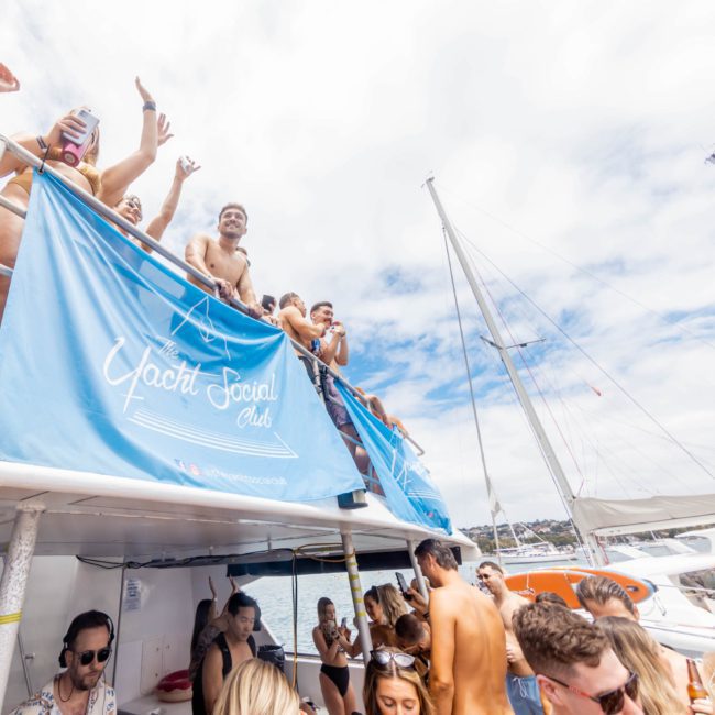 A group of people enjoys a sunny day on a private yacht charter Sydney Harbour decorated with a "Yacht Social Club" banner. Some are standing on the upper deck, while others are below. Sails and boats are visible in the background, perfect for a memorable Sydney boat party hire.