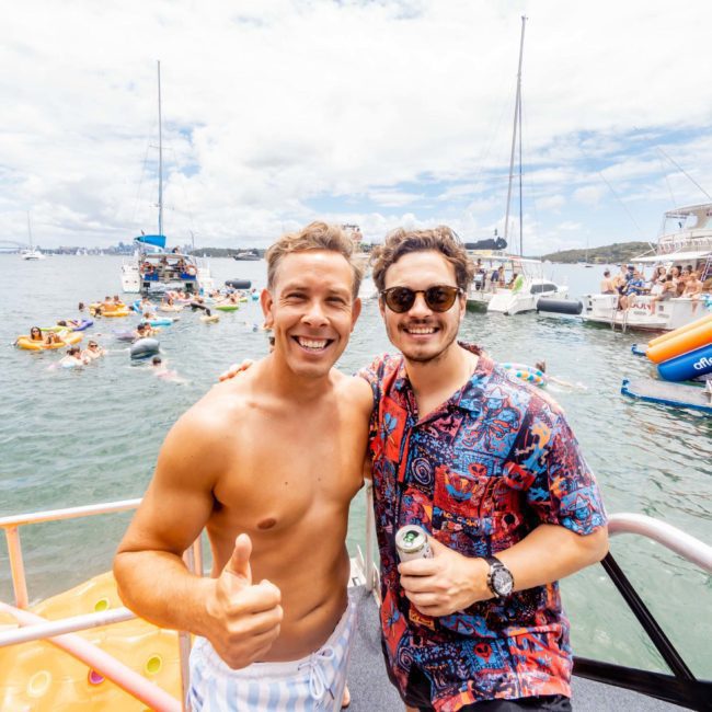 Two men stand on a boat deck surrounded by other boats and floats. One man is shirtless and giving a thumbs up, while the other wears a patterned shirt and sunglasses. Both are smiling at the camera, enjoying their day at a lively Sydney boat party hire.
