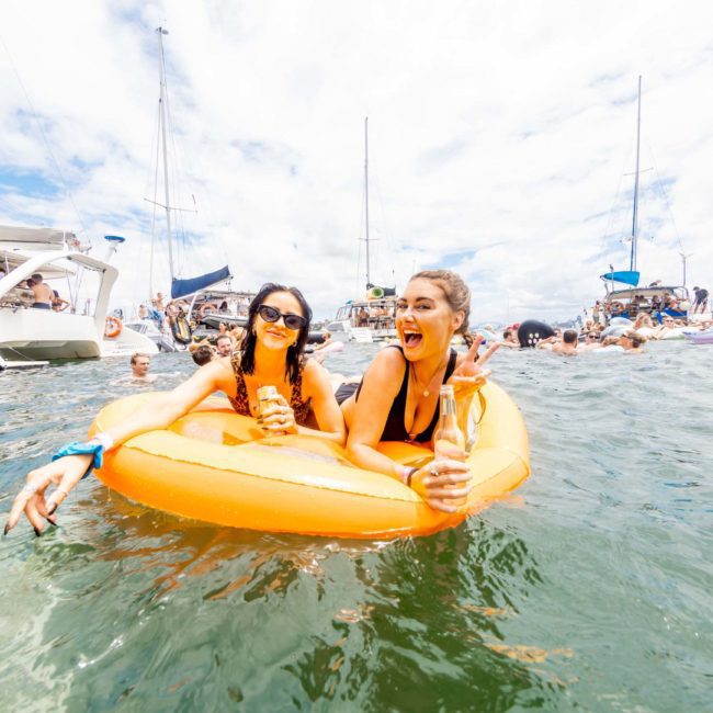 Two women on an inflatable float in the water, holding drinks, surrounded by other people and boats in the background under a bright sky, enjoying a private yacht charter Sydney Harbour.