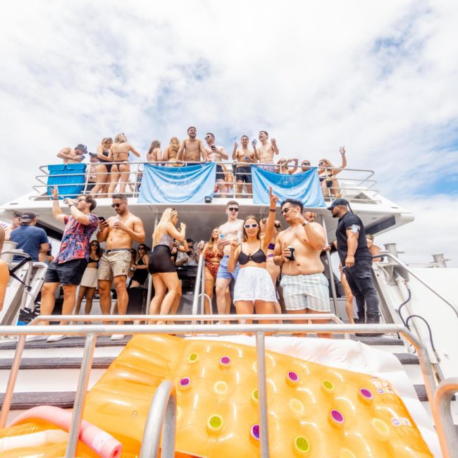 People enjoying a party on a private yacht charter in Sydney Harbour, with many individuals on the upper deck and some holding drinks. An inflatable toy is visible at the bottom. The sky is partly cloudy.