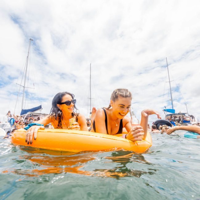 Two women on an orange float enjoy a sunny day in the water, surrounded by other people and boats during a lively Catamaran party in Sydney.