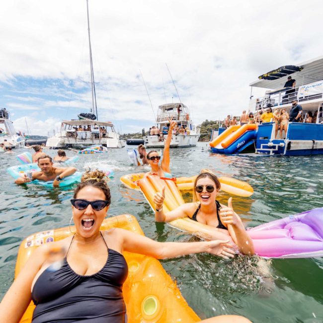 Group of people on inflatable floats having fun in the water, surrounded by boats with slides, under a partly cloudy sky—a perfect setting for a lively Sydney boat party hire or private yacht charter.
