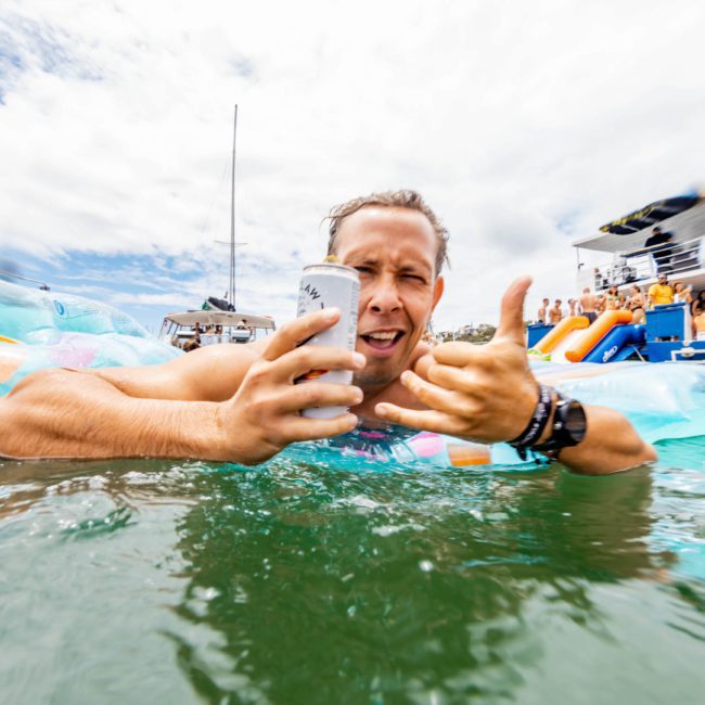 A person holding a drink in one hand and making a hand gesture with the other floats on an inflatable in the water. Boats and people enjoy a perfect day in the background under a partly cloudy sky, reminiscent of fun-filled Sydney boat party hires.
