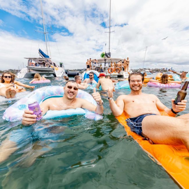 People relax and float on inflatables in the water, holding drinks, with boats and other people in the background on a sunny day during a luxury yacht hire Sydney.
