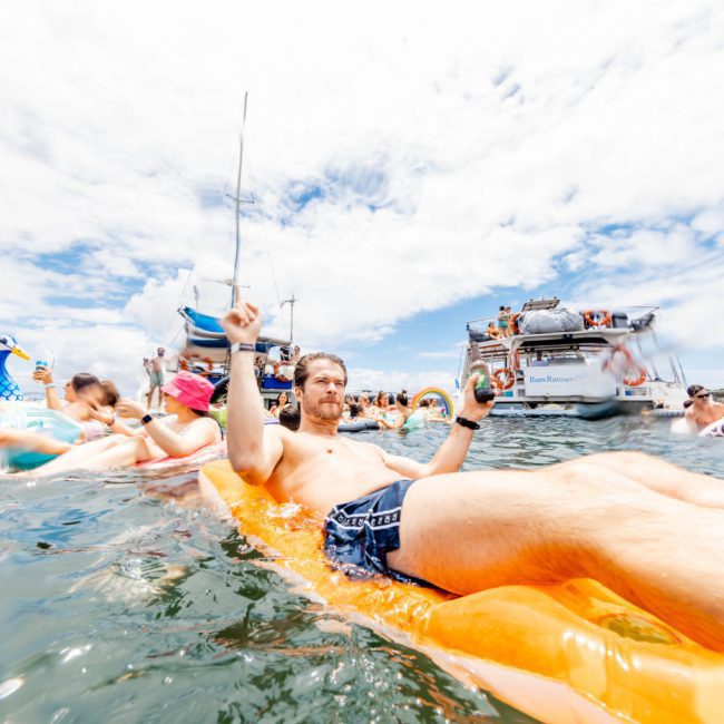 A man relaxes on an orange float in the water, surrounded by other people on inflatables. Boats and a partly cloudy sky are visible in the background, perfect for a private yacht charter Sydney Harbour experience.