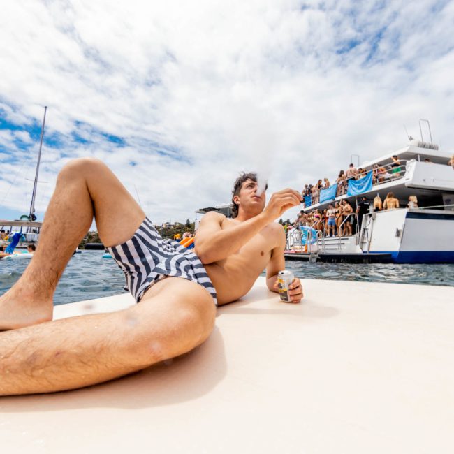 Man in swim trunks lounges on a raft holding a drink, with a DJ boat hire Sydney in the background and people in swimwear enjoying the water.