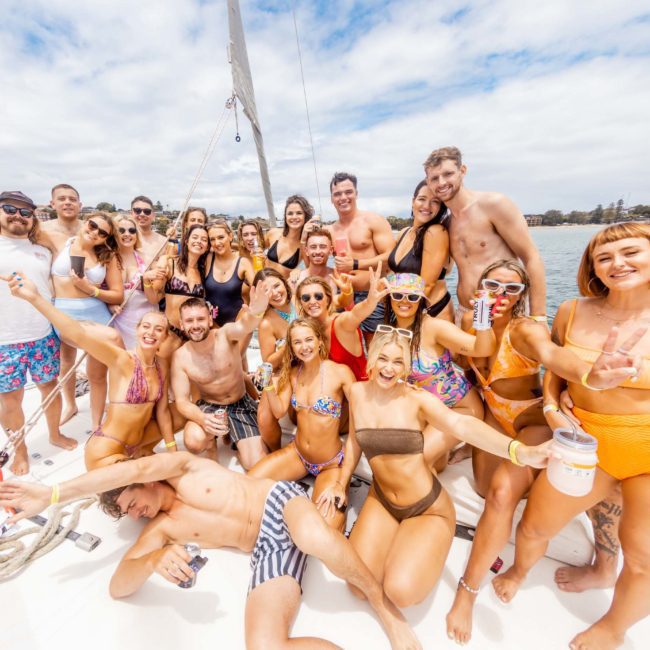 A group of people in swimsuits is gathered on a boat, smiling and posing for a photo. The luxury yacht is on water with other vessels in the background under a partly cloudy sky.