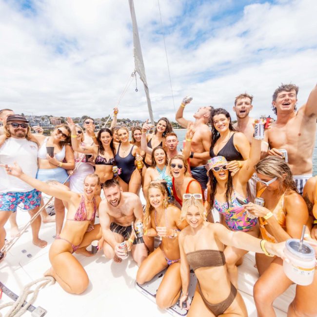 A large group of people in swimwear are gathered on a boat, smiling and raising their arms. Some are holding drinks. It is a sunny day with a cloudy sky, and another boat is visible in the background—perfect for a lively Sydney boat party hire or corporate boat events Sydney.