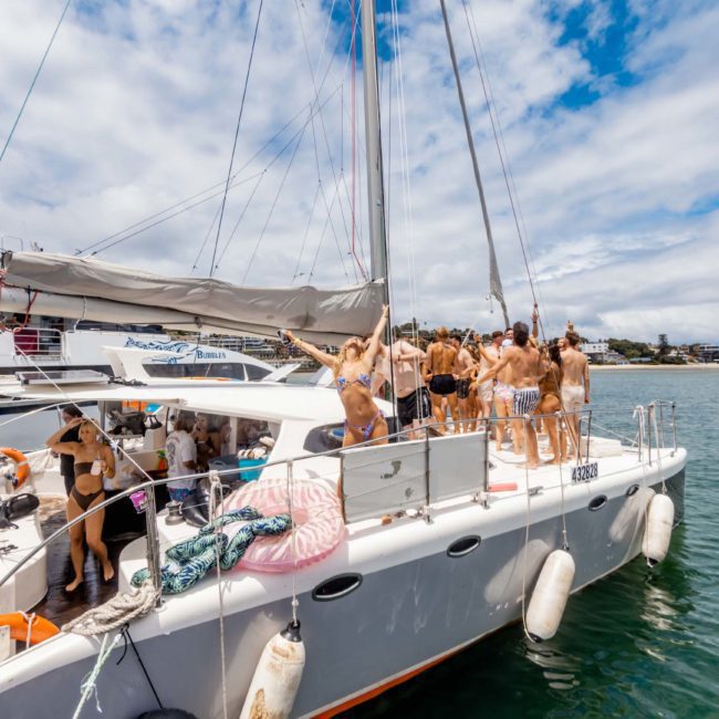 A group of people stands and sits on a catamaran sailboat in the water under a partly cloudy sky, with another boat visible in the background. Some are wearing swimwear, enjoying what looks like a lively Sydney boat party hire.
