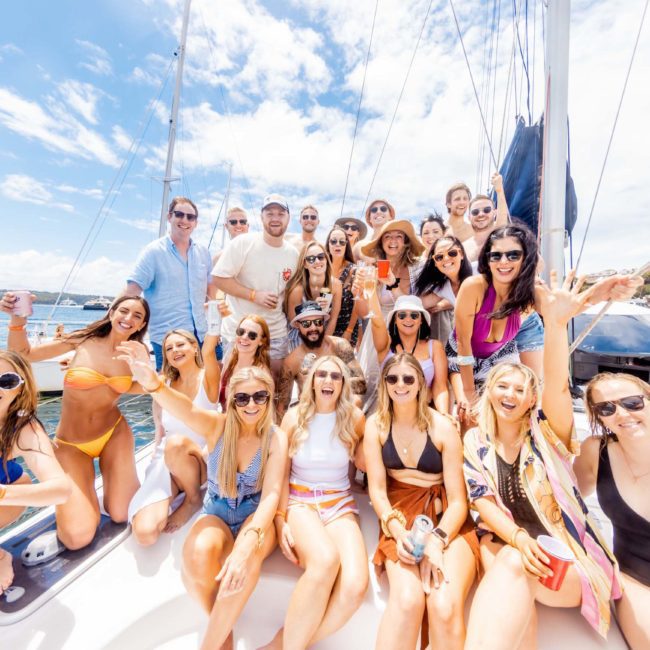 A group of young adults in swimwear and casual attire, smiling and posing on a sailboat under a sunny sky. Some hold drinks as they enjoy a Sydney boat party hire at the marina, with other boats in the background.