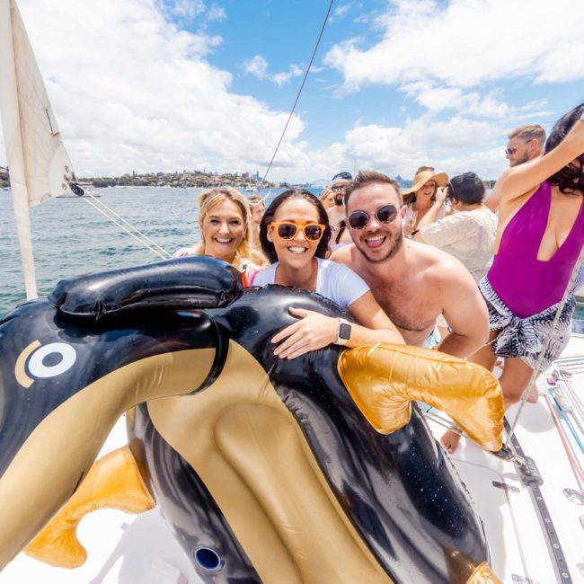 A group of people aboard a sailboat, smiling and posing with a large inflatable penguin. The weather is sunny, and the background shows a waterfront landscape perfect for a corporate boat event in Sydney.