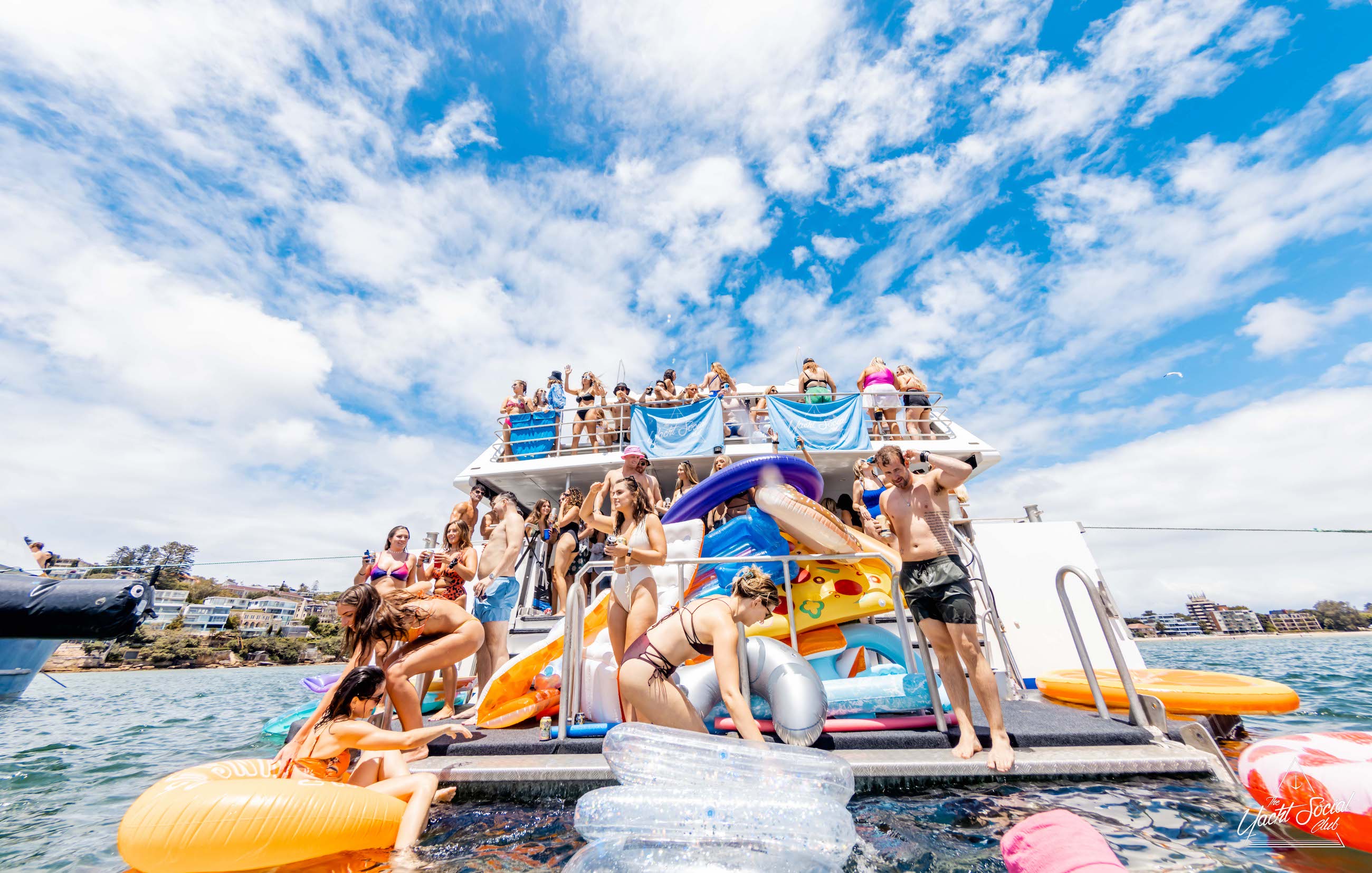 A large group of people in swimwear is having a fantastic time on a double-deck boat with slides, inflatables, and floatations in the water, under a bright sunny sky with scattered clouds. Perfect for corporate boat events Sydney or Sydney boat party hire.