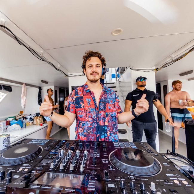 A person wearing headphones stands behind a DJ mixer on a boat in Sydney. Other people in swimsuits are in the background, some talking and some lounging around, enjoying the Catamaran party Sydney offers.