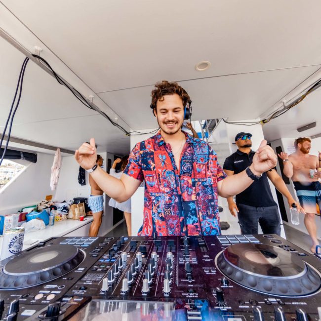A person stands behind DJ equipment on a boat, wearing headphones, giving thumbs up. Other people are in the background, enjoying the vibrant atmosphere of a Catamaran party in Sydney.