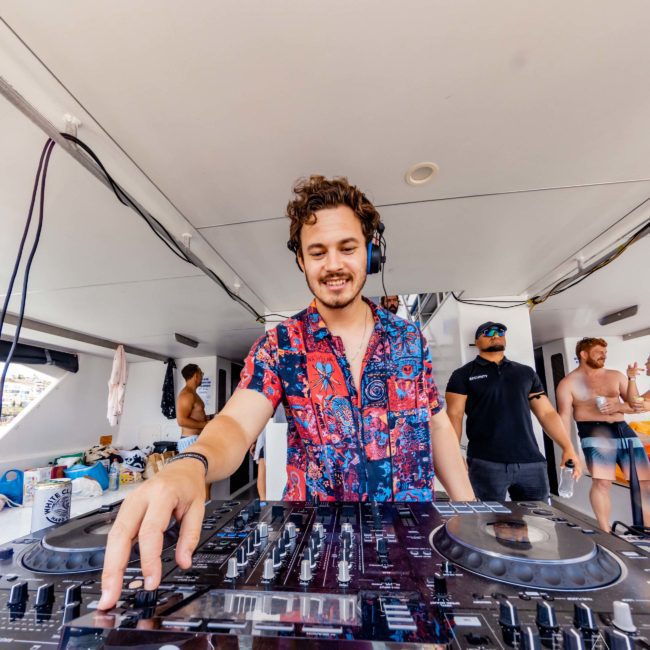 A DJ wearing a colorful shirt operates a Pioneer DJ mixer on a yacht, with people in casual attire in the background, highlighting the vibrant atmosphere of Sydney boat party hire.
